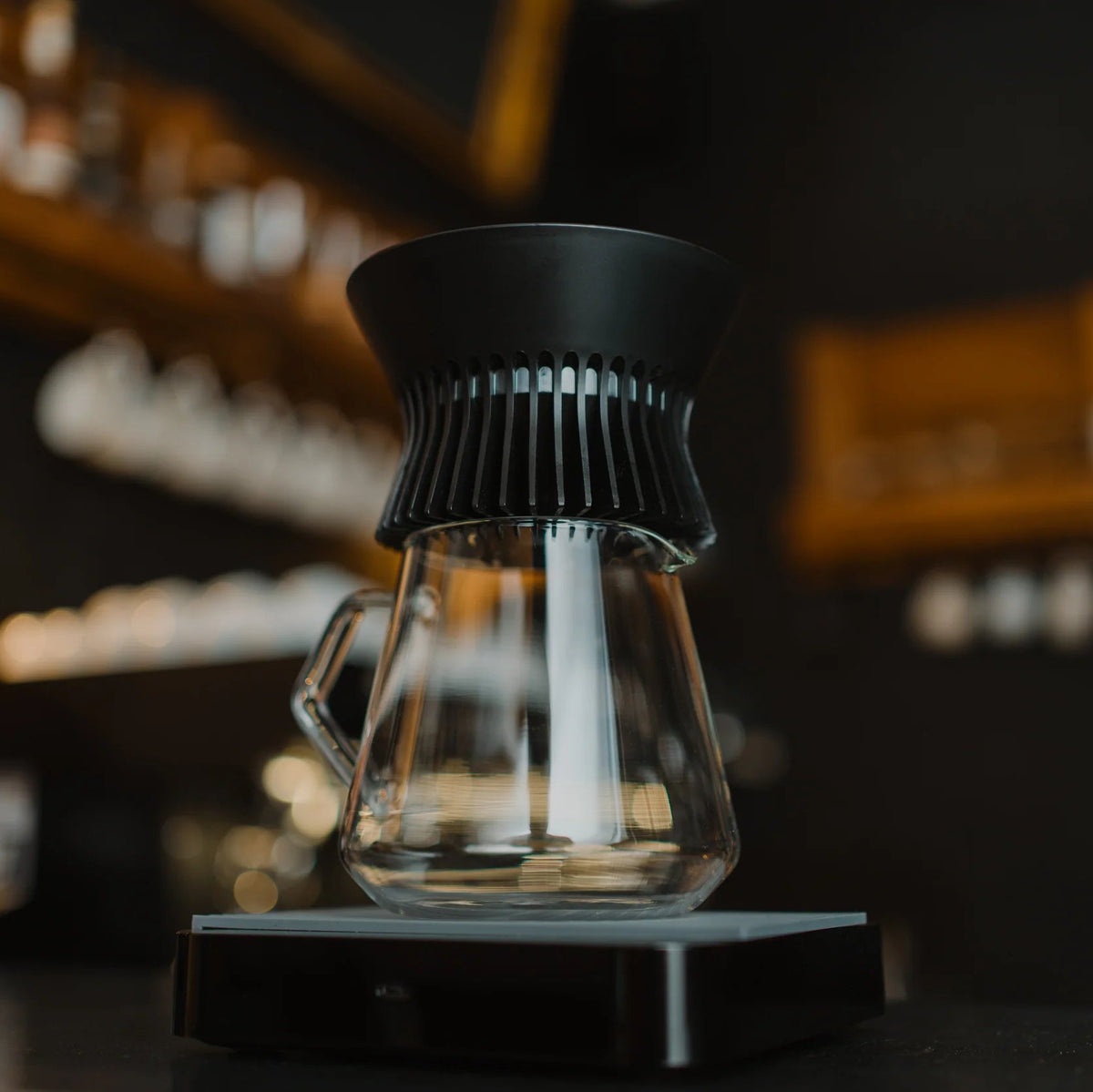 A glass pour-over coffee maker with a black conical filter sits on a digital scale on a dark countertop, with a blurred bar or kitchen background.
